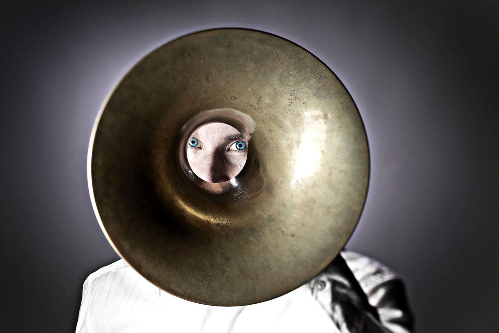 A worn brass instrument bell held in front of a person's face, eyes showing through the small opening.