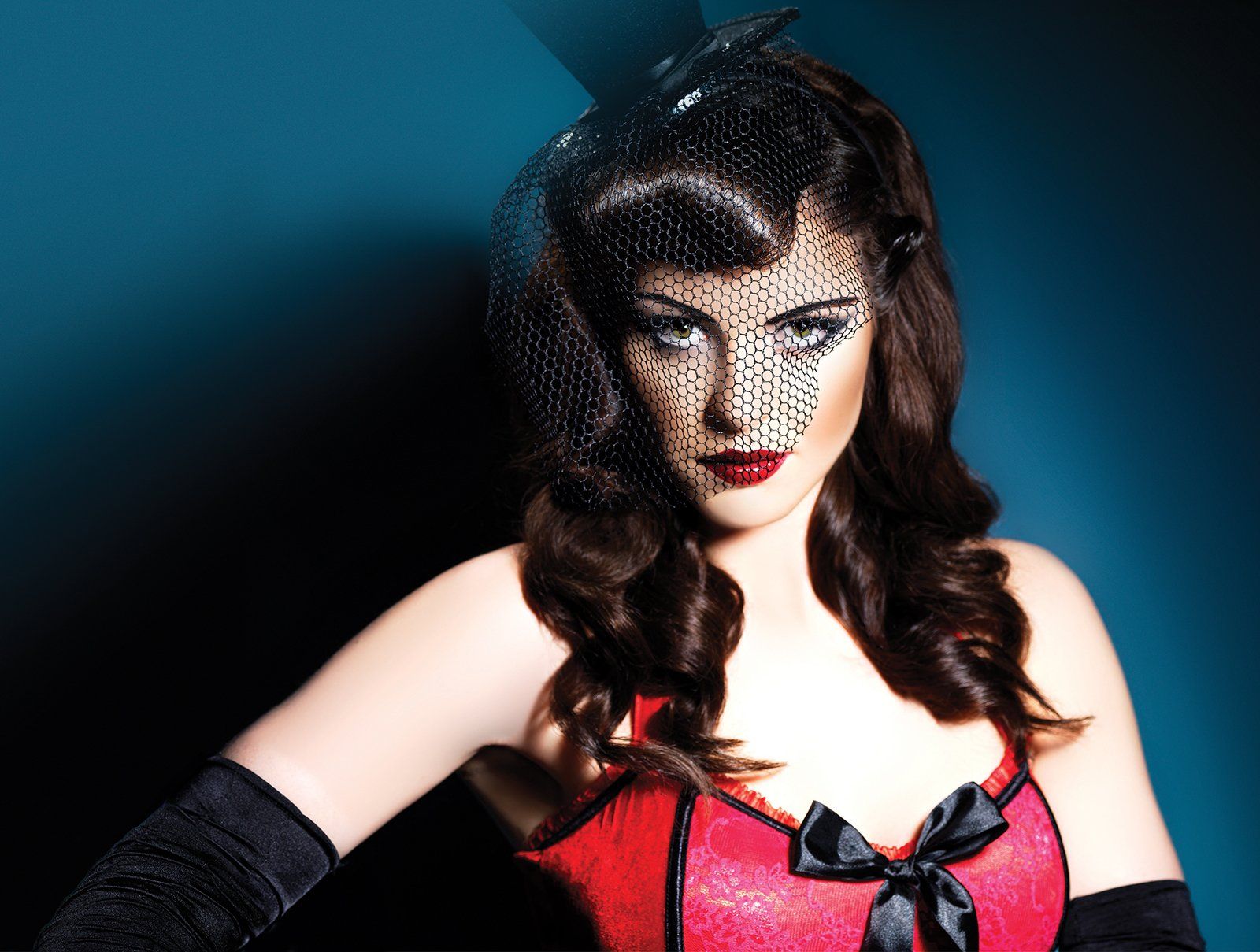 A woman in a black net birdcage veil, small top hat, long gloves and a red corset, against a teal background.